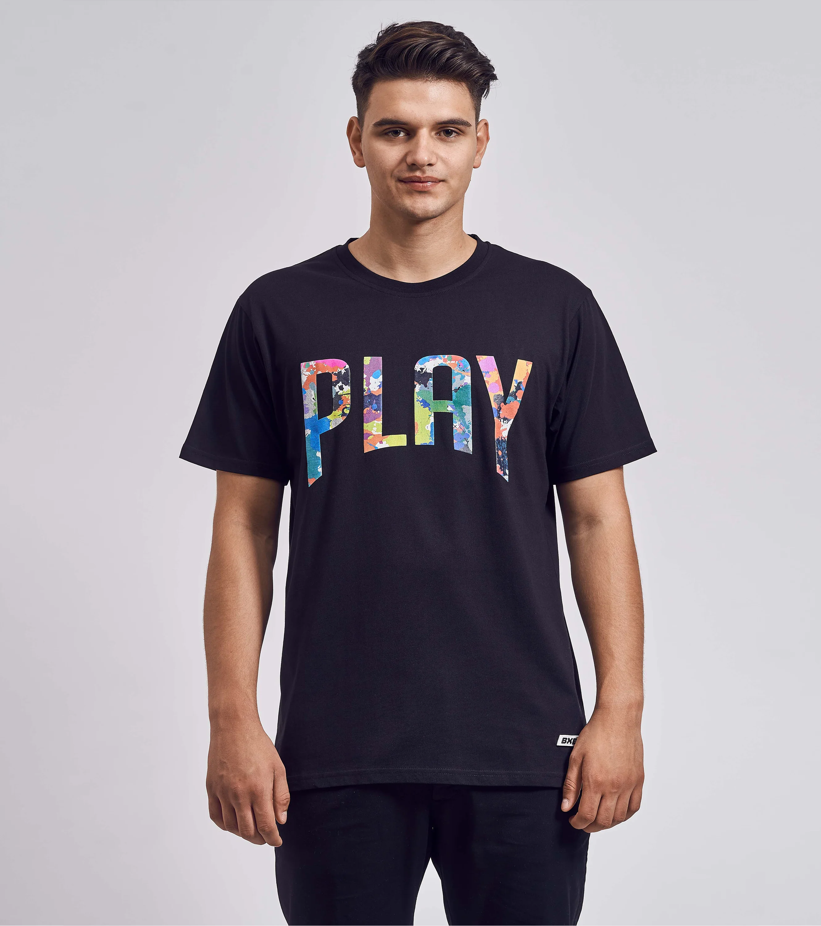 Schwarzes T-Shirt mit buntem „PLAY“-Schriftzug, getragen von männlichem Model, fotografiert von vorn vor grauem Hintergrund