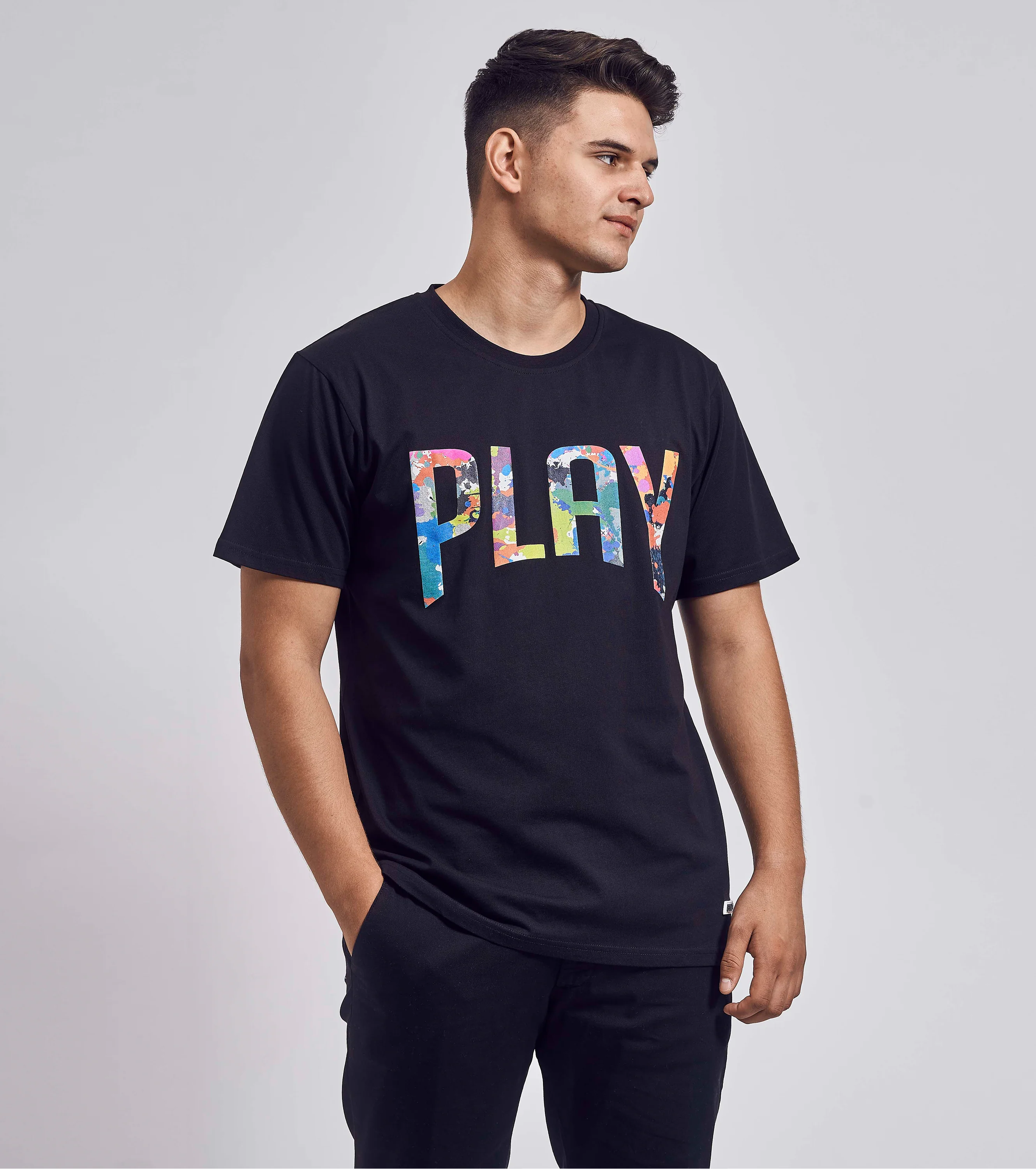 Schwarzes T-Shirt mit buntem „PLAY“-Schriftzug, getragen von männlichem Model in seitlicher Pose mit Blick nach rechts