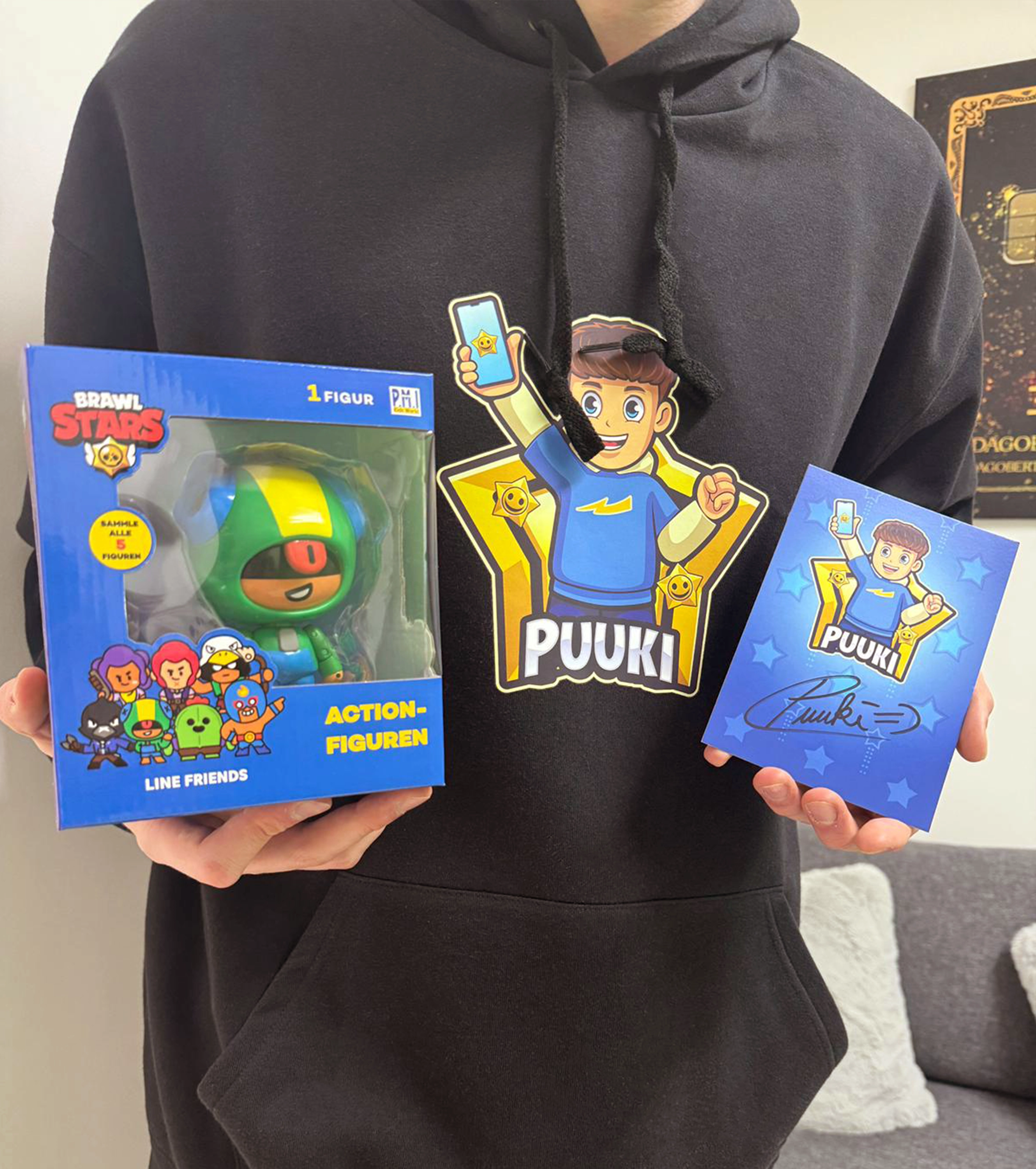 Nahaufnahme des Puuki-Merch-Bundles: schwarzer Hoodie mit Puuki-Comicdruck, signierte Autogrammkarte und bunte Brawl Stars Action-Figur – perfekt fuer junge YouTube-Fans und Sammler.