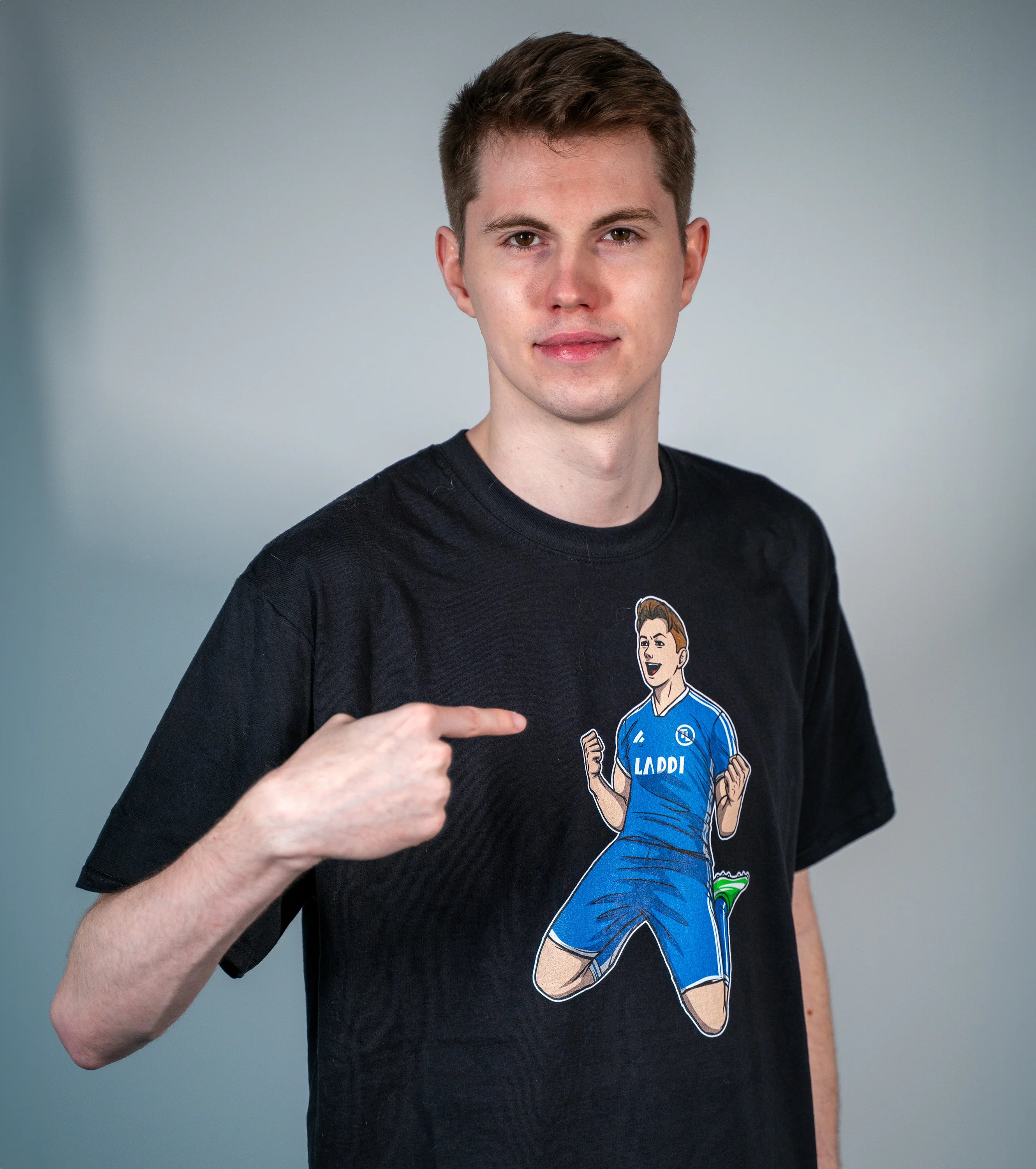 Tim Latka trägt ein schwarzes T-Shirt mit Comic-Motiv seiner jubelnden Figur im blauen Trikot und zeigt auf das Laddi-Design auf seiner Brust.

